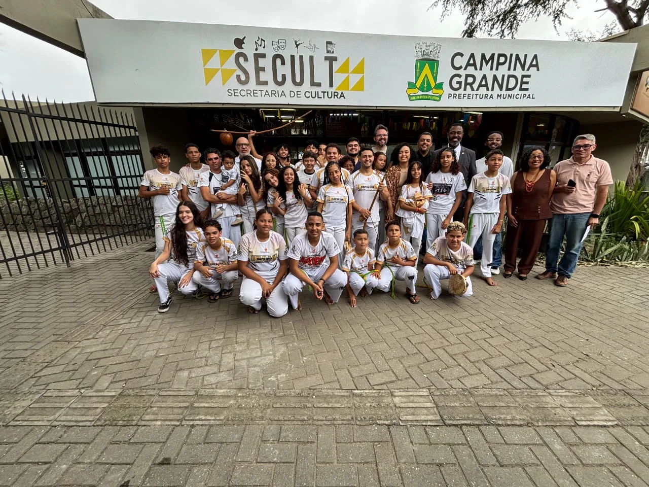 Grupo de crianças e adolescentes com uniforme branco de capoeira posa com autoridades na entrada da Secretaria de Cultura de Campina Grande.