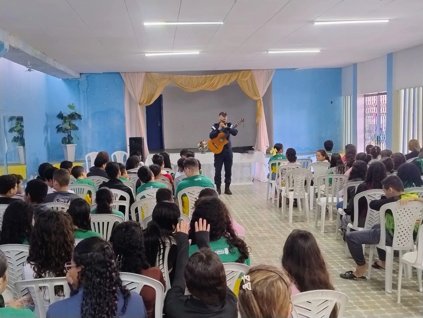 Agente da Guarda Civil Municipal toca violão sobre um pequeno palco de auditório escolar, diante de estudantes de uniforme sentados em cadeiras.