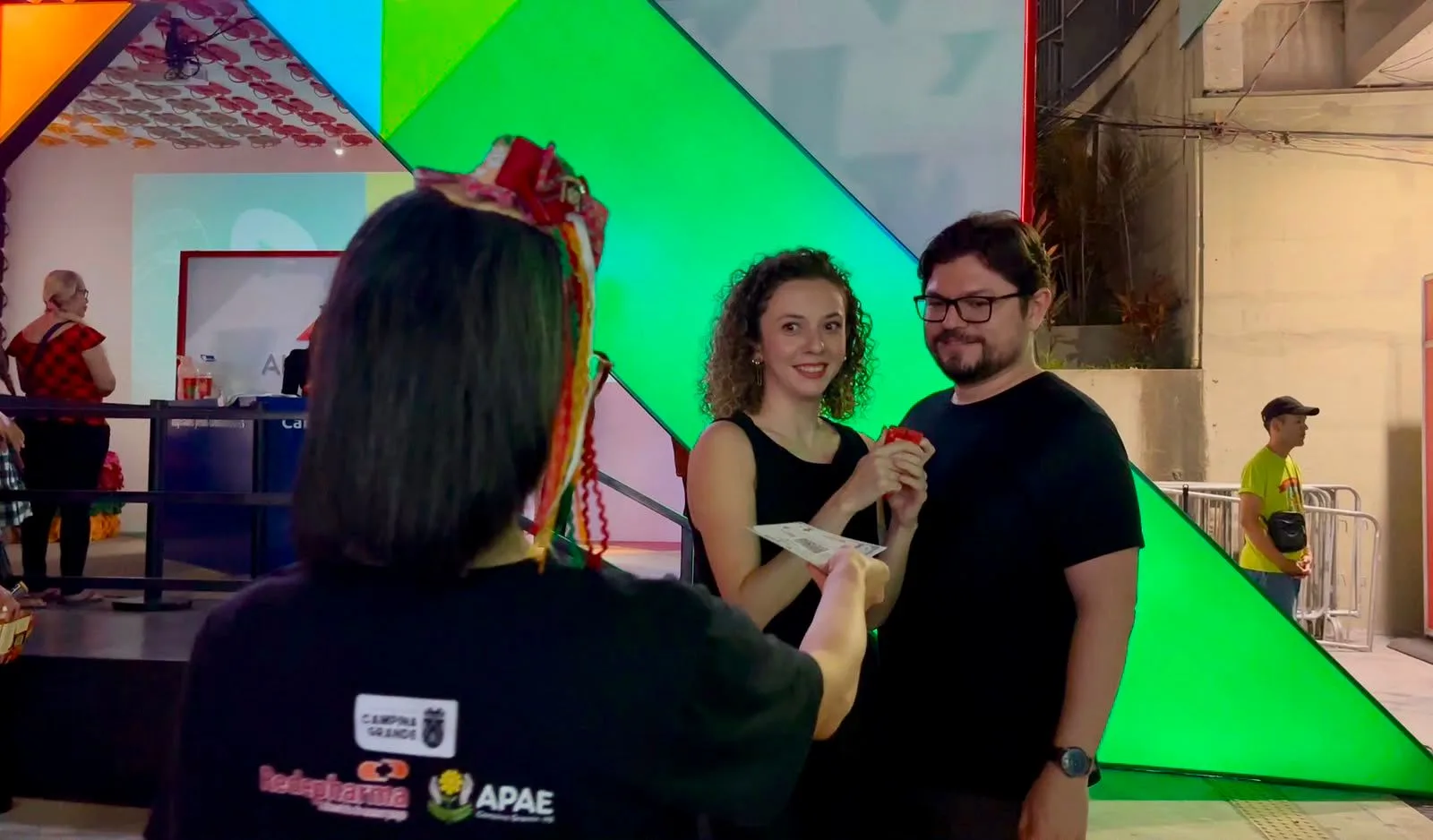 Atendente de camiseta preta com logos da Prefeitura, Redepharma e Apae entrega um folheto a um casal de visitantes, sob iluminação junina.