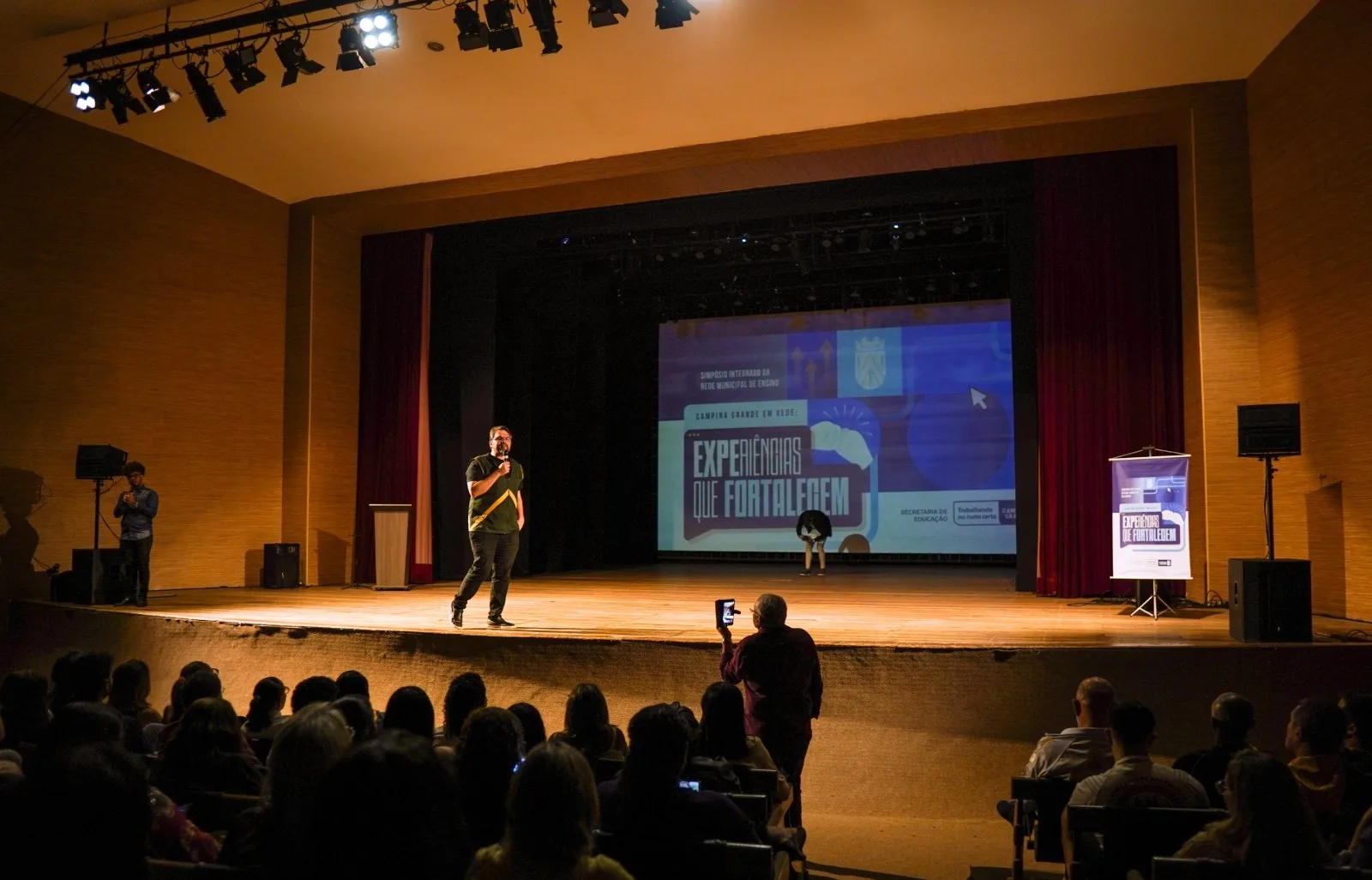 Homem fala ao microfone no palco do teatro diante da plateia, com telão exibindo a marca do simpósio ao fundo.